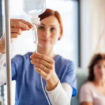 A front view of nurse with IV drip and patient in bed in hospital room.